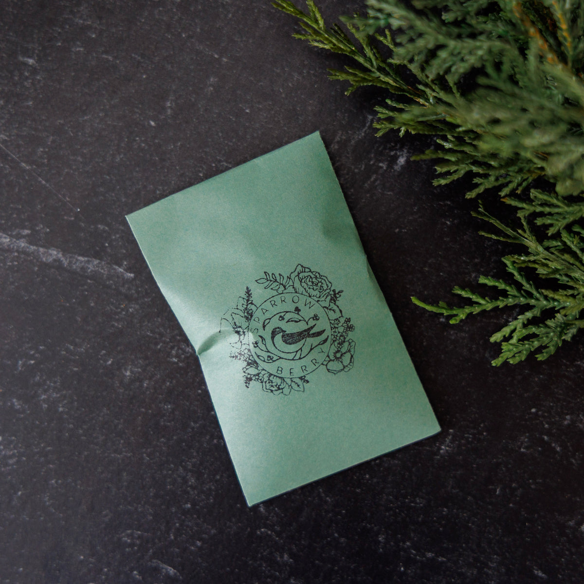 A mint green paper envelope with a black printed logo of a fish surrounded by flowers and the words 'SPARROW BERRY', placed on a dark textured surface next to green foliage.