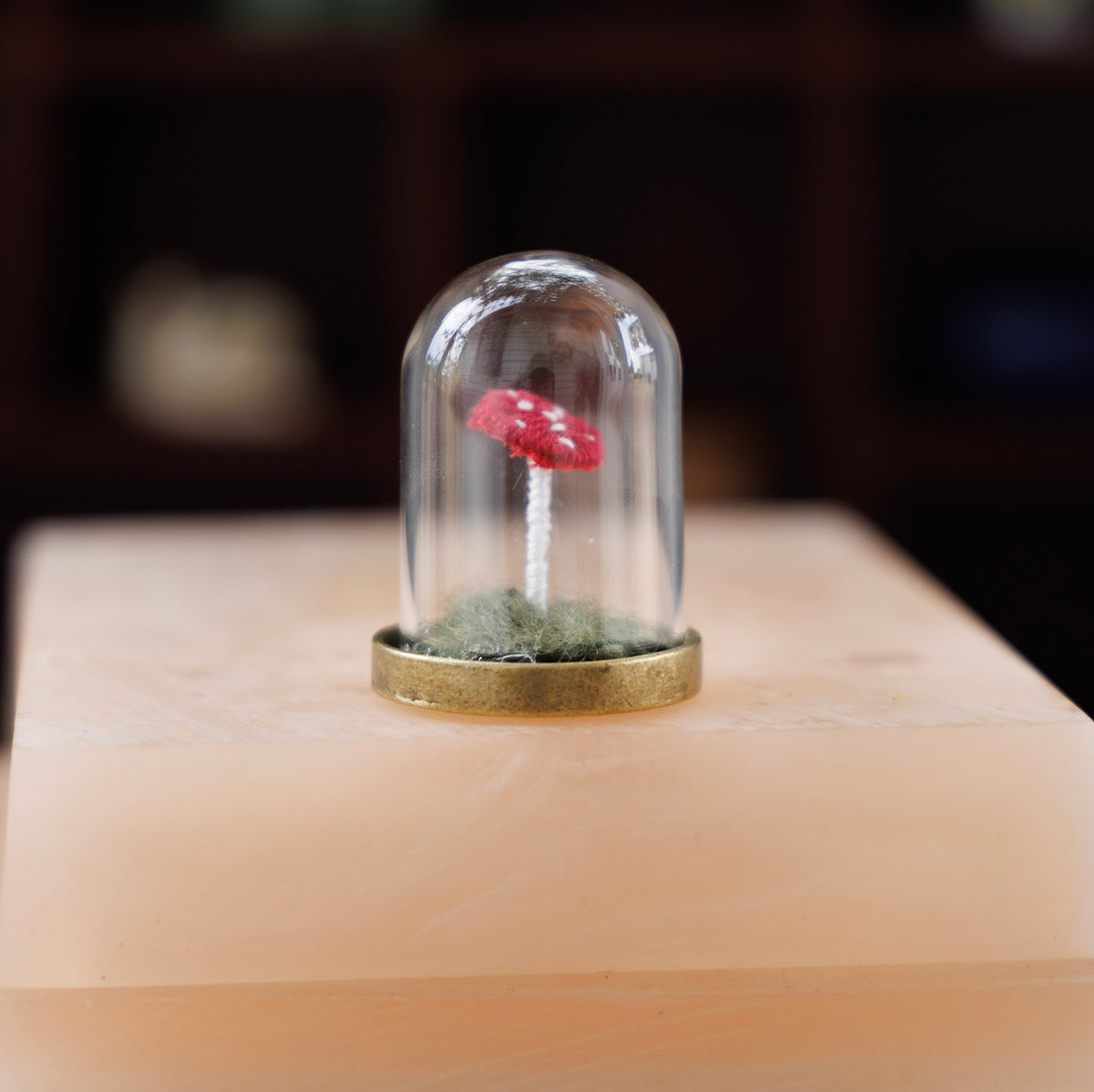 A miniature red and white spotted mushroom under a clear glass dome with a gold metal base, set on a pale peach stone slab against a dark blurred background.