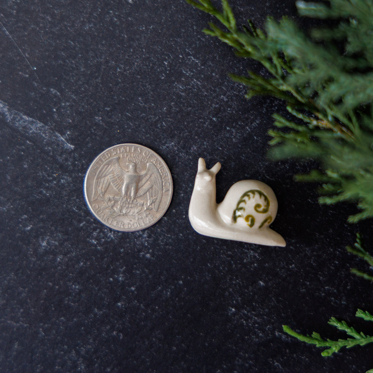 A small cream-colored ceramic snail figurine with green leaf accents on its shell, shown next to a US quarter coin for scale on a dark textured surface with green foliage in the corner.
