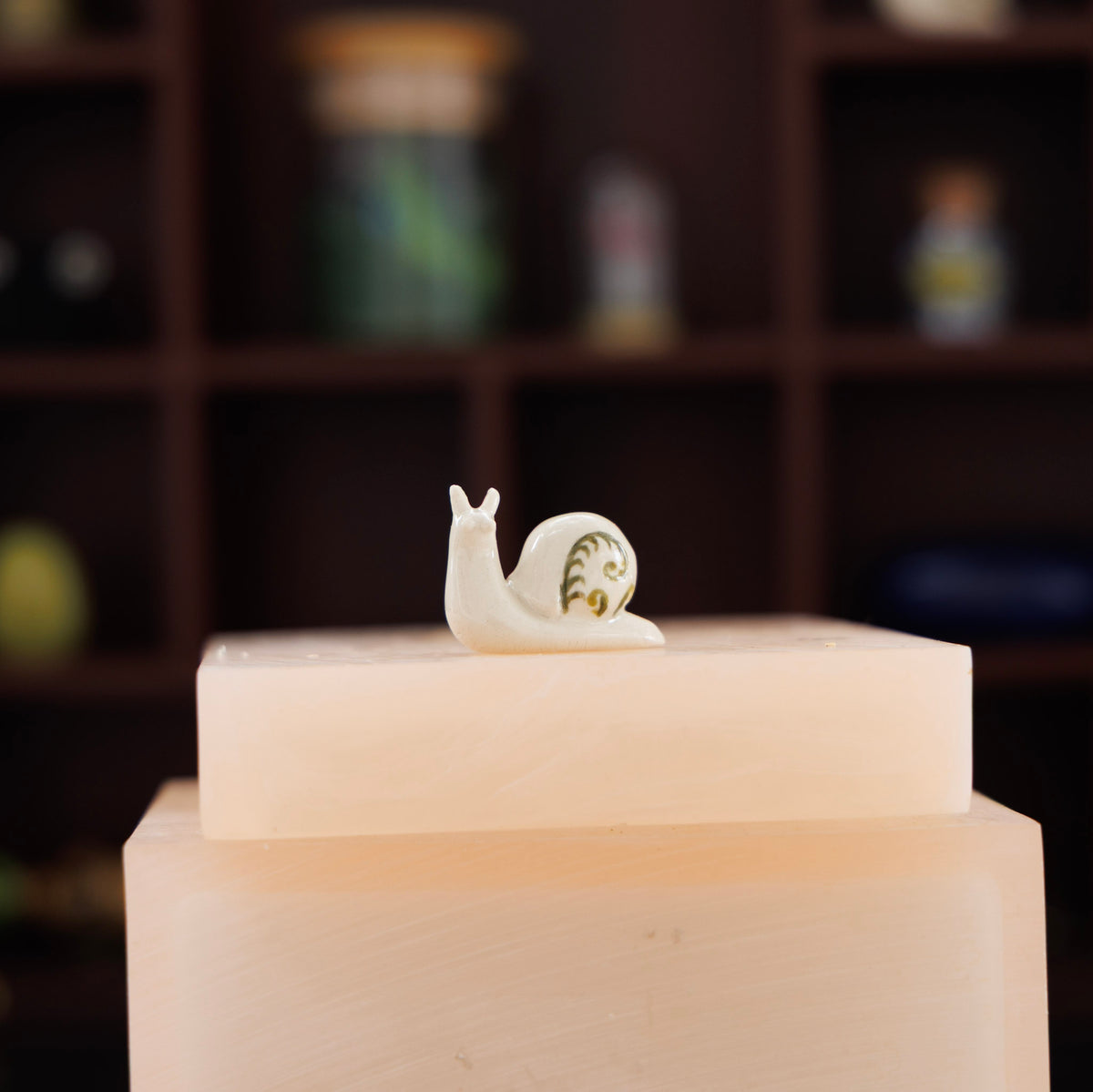 A small white snail figurine with a green and gold shell detail, displayed on top of two stacked pale pink blocks against a blurred dark brown shelving background.