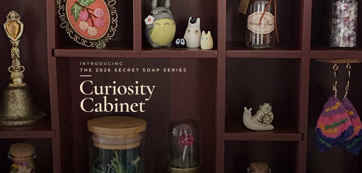 A promotional graphic for 'The 2026 Secret Soap Series: Curiosity Cabinet' showing a dark brown wooden shelf filled with various decorative soaps, jars, and small figurines.