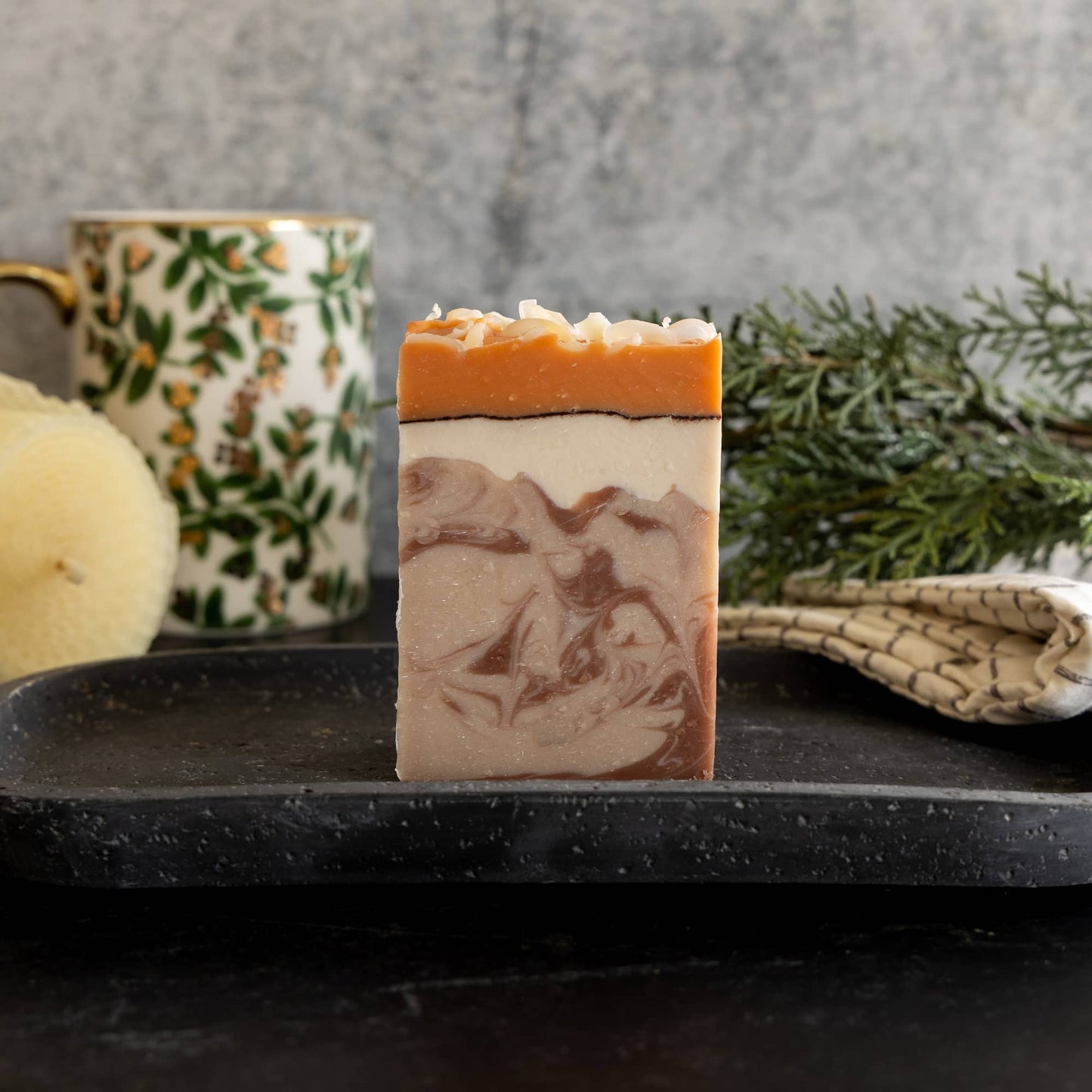 Bar of soap with a natural design on a black surface, with a mug and greenery in the background.