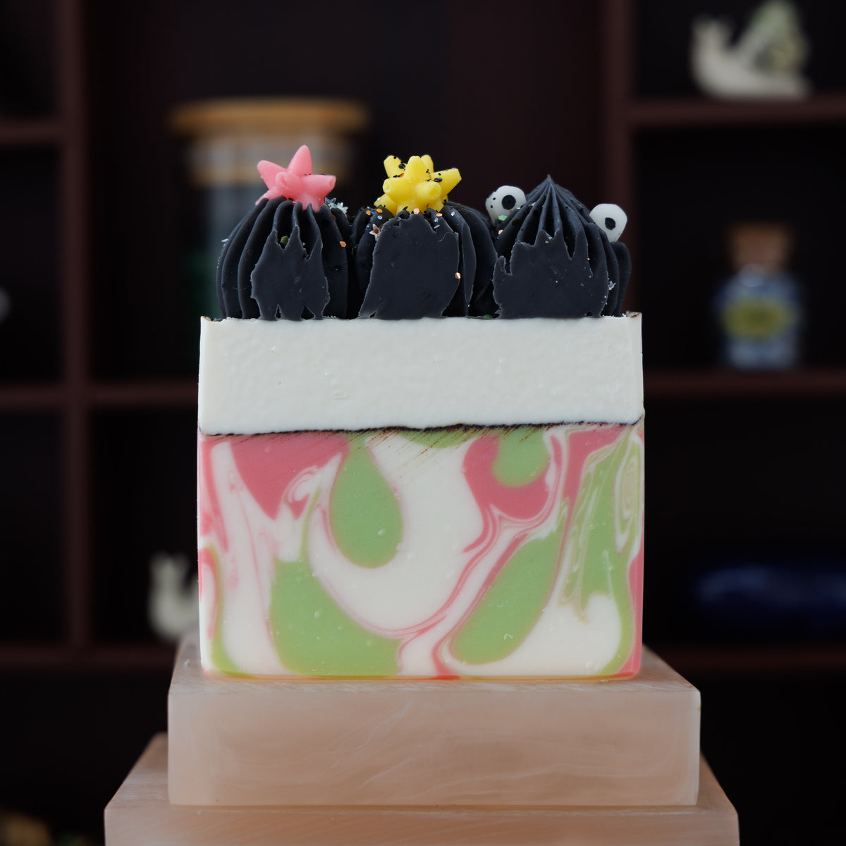 A decorative rectangular soap bar with a pink, green, and white marbled pattern, topped with three black piped shapes and small star accents, displayed on a pink stone pedestal against a dark wood shelf background.