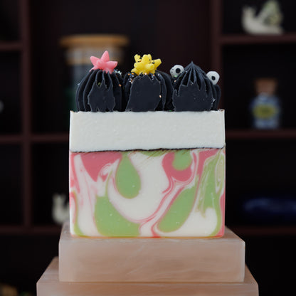 A decorative rectangular soap bar with a pink, green, and white marbled pattern, topped with three black piped shapes and small star accents, displayed on a pink stone pedestal against a dark wood shelf background.