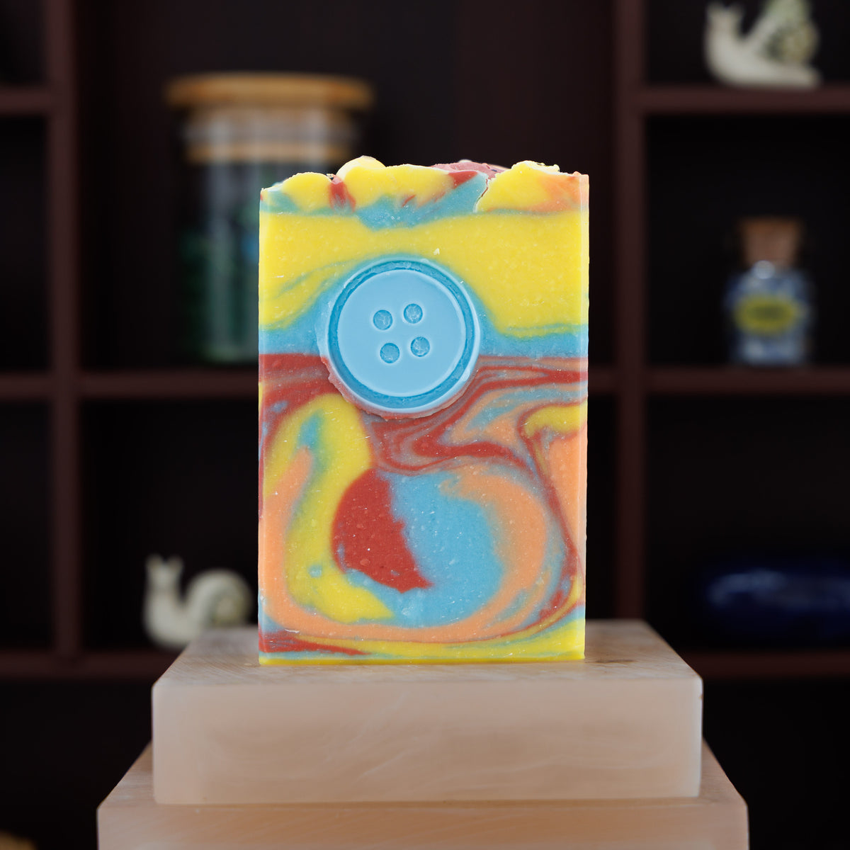 A handmade rectangular soap bar with a marbled swirl pattern of yellow, blue, orange, and red, featuring a light blue button detail on the front, displayed on a stone block against a dark wooden shelf background.