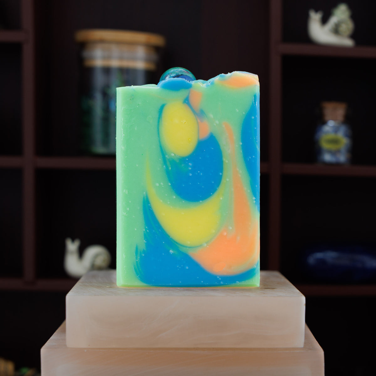 A handmade rectangular soap bar with a marbled swirl pattern of green, blue, yellow, and orange, topped with a glass marble, displayed on a stone block against a dark wooden shelf background.