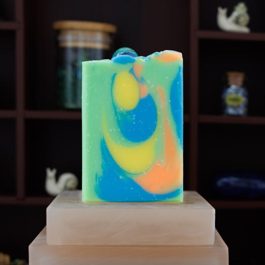 A handmade rectangular soap bar with a marbled swirl pattern of green, blue, yellow, and orange, topped with a glass marble, displayed on a stone block against a dark wooden shelf background.