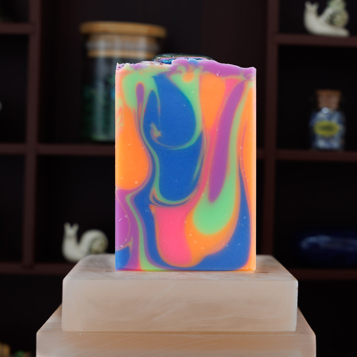 A rectangular handmade soap bar with a vibrant marbled swirl pattern of blue, purple, orange, and green, displayed on a stone block against a dark wooden shelf background.