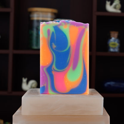 A rectangular handmade soap bar with a vibrant marbled swirl pattern of blue, purple, orange, and green, displayed on a stone block against a dark wooden shelf background.