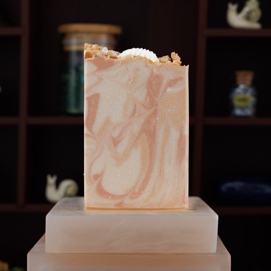 A rectangular handmade soap bar with a peach and cream marbled pattern, topped with a white seashell, displayed on a light pink stone pedestal against a dark wooden shelf background.